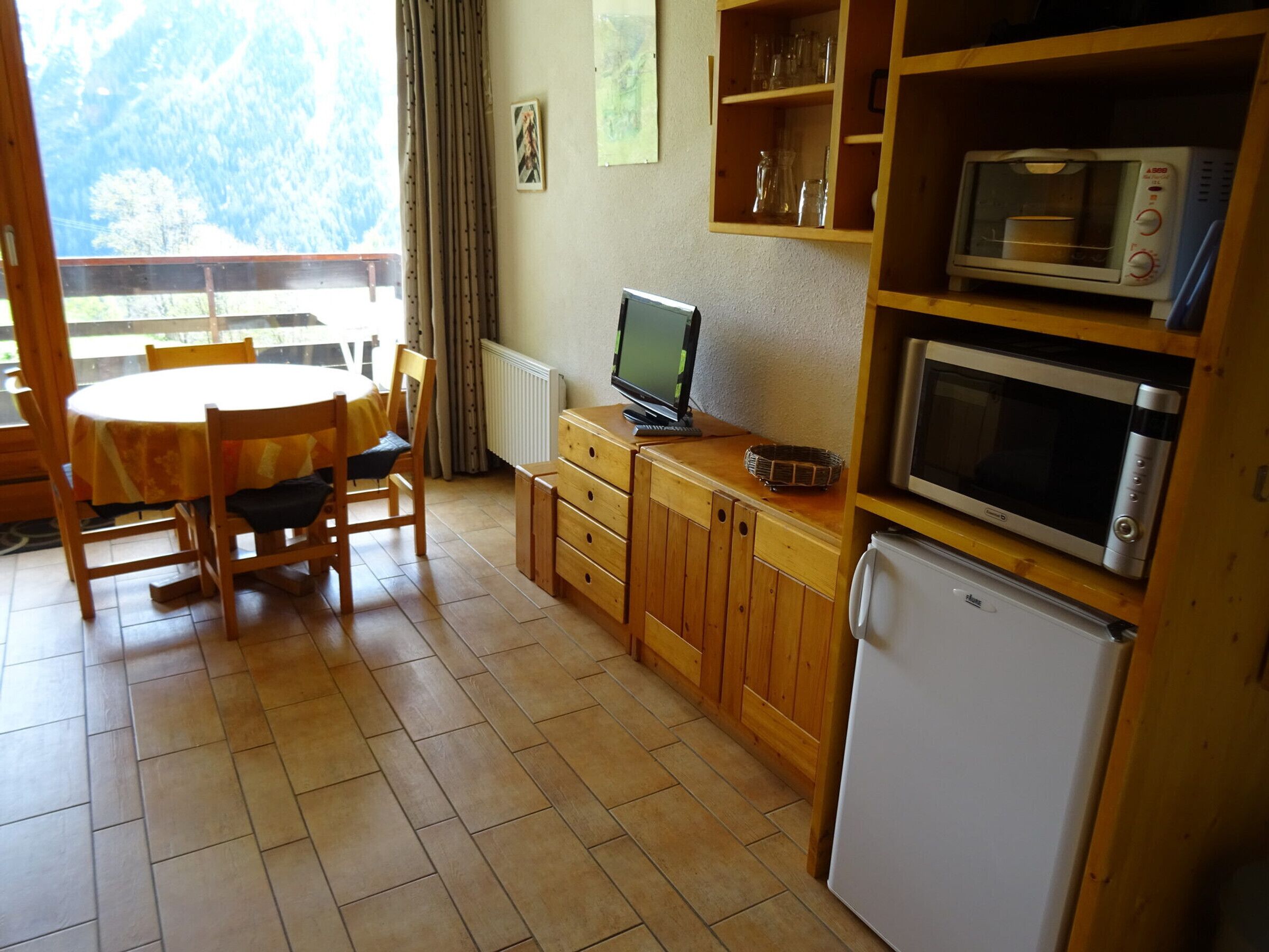 A compact kitchen with wooden cabinets, a microwave, a refrigerator, and a dining area with a table and chairs.