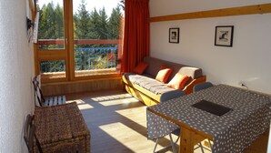 Ein gemütliches Zimmer mit einer Couch, einem Esstisch und Blick auf Bäume durch große Fenster.