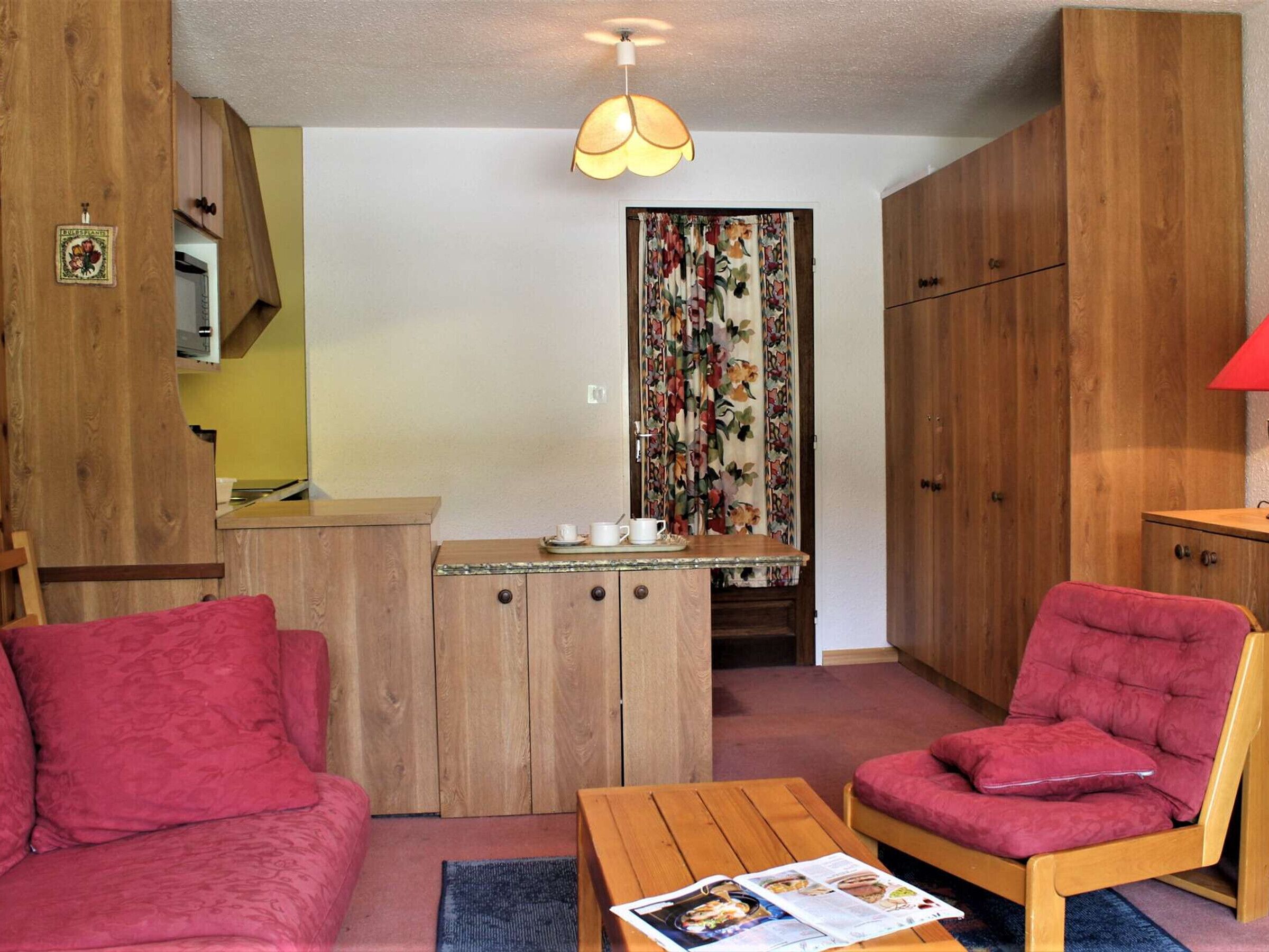 Een compacte leefruimte met een kitchenette, een eettafel, twee rode fauteuils en een plafondlamp.