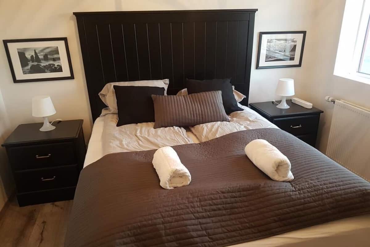 Un lit bien fait, recouvert d’une couverture marron, avec deux oreillers et deux serviettes enroulées posées dessus. Il y a deux tables de chevet, chacune avec une lampe et un tableau encadré. La pièce a un plancher en bois et une fenêtre sur le côté droit.