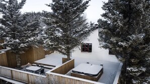 Una terraza de madera con un jacuzzi y una mesa, rodeada de árboles cubiertos de nieve.