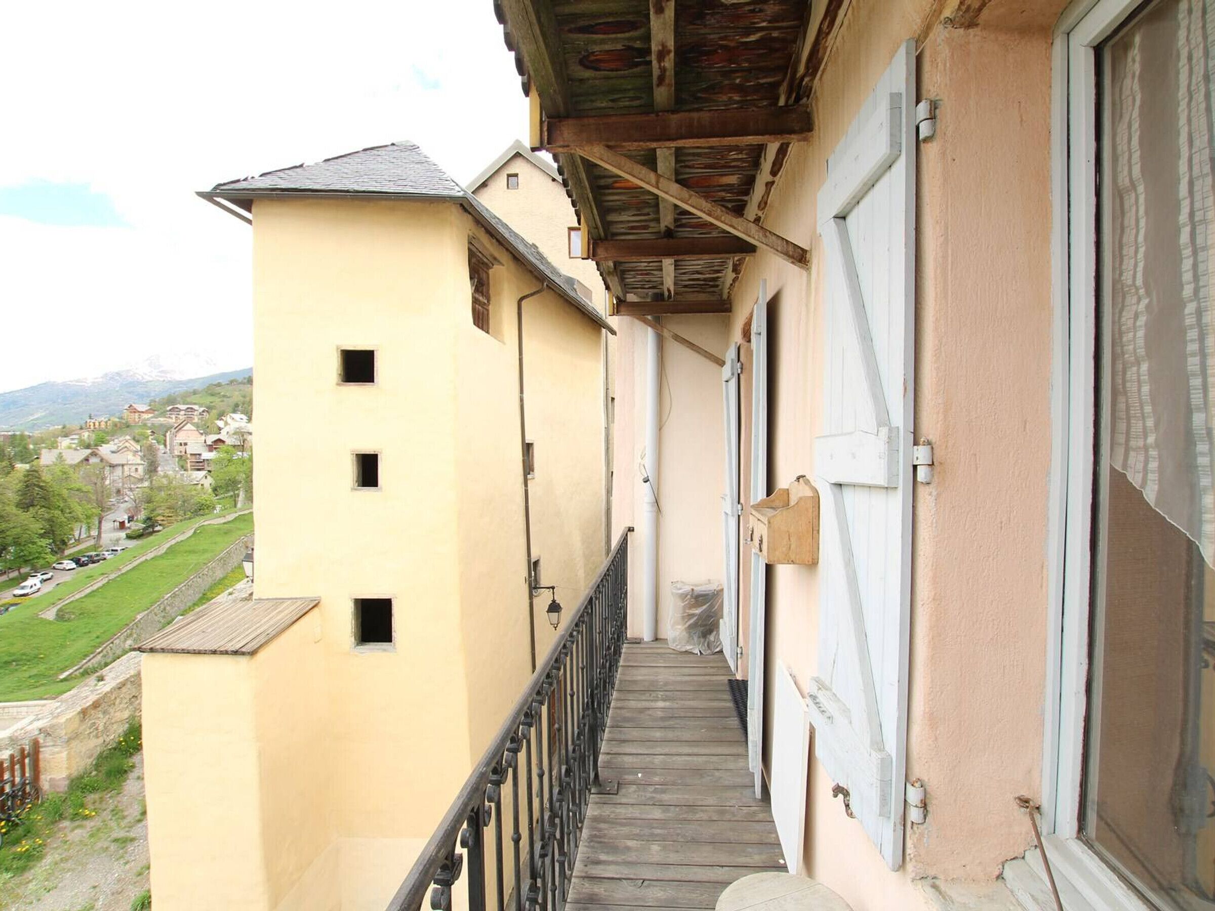 Un balcone stretto con ringhiera in legno, un edificio beige con molte finestre e vista su un paesaggio verde e edifici in lontananza.