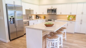 A modern kitchen with white cabinets, stainless steel appliances, a central island with stools, and a wooden floor. - Ma'alaea Surf, Unit E1-2 BR Ground Floor Beachfront Condo, AC, Wifi, Pool (Kihei)