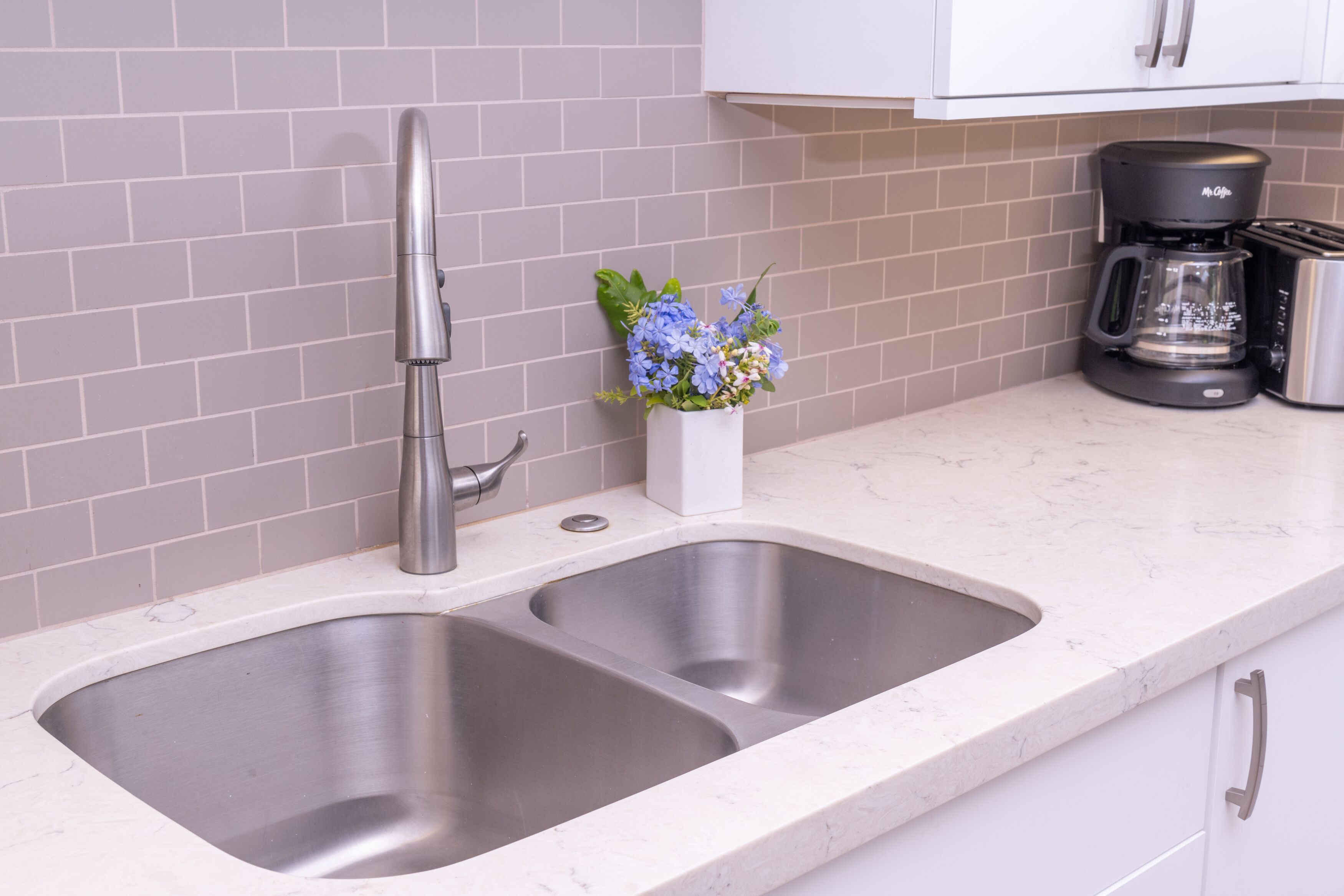 A kitchen with a double stainless steel sink, a modern faucet, a coffee maker, and a small vase with flowers on the countertop.