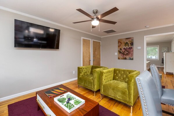 Un salon équipé d’un téléviseur à écran plat, d’un ventilateur de plafond, de deux fauteuils verts, d’une table basse en bois et d’une chaise bleue.