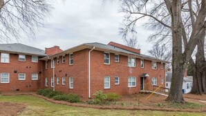 A multi-story brick building with a flat roof, multiple windows, and a small front yard with a tree and shrubs. - 1BR Sparkling Clean Apartment, KING Bed (Winston-Salem)