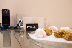 A table with two white coffee cups, a sign reading "Prati 25.7 Suites in Rome," and a glass dish with pastries.
