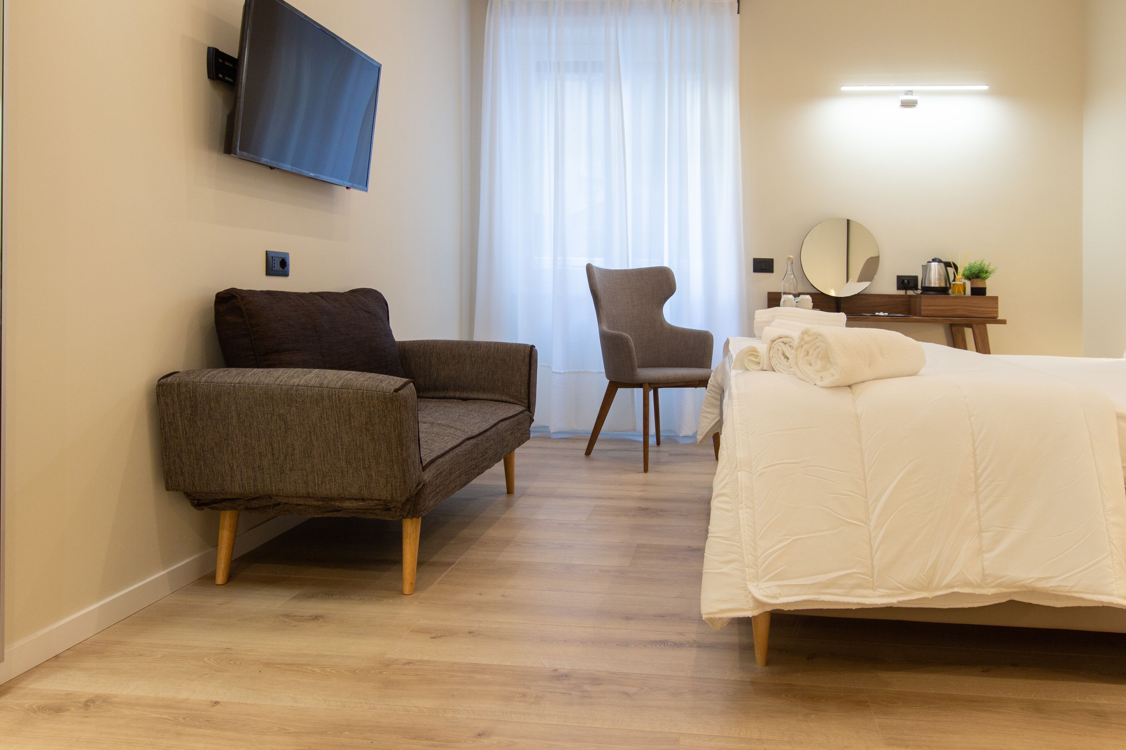 A hotel room with a sofa, a chair, a bed, a desk with a mirror, and a TV mounted on the wall.