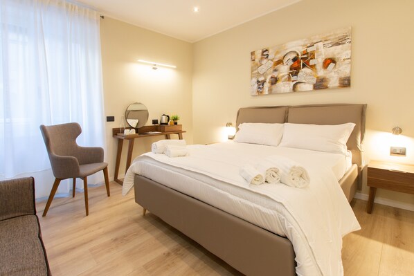 A hotel room with a large bed, a chair, a desk with a mirror, and a wall-mounted artwork.
