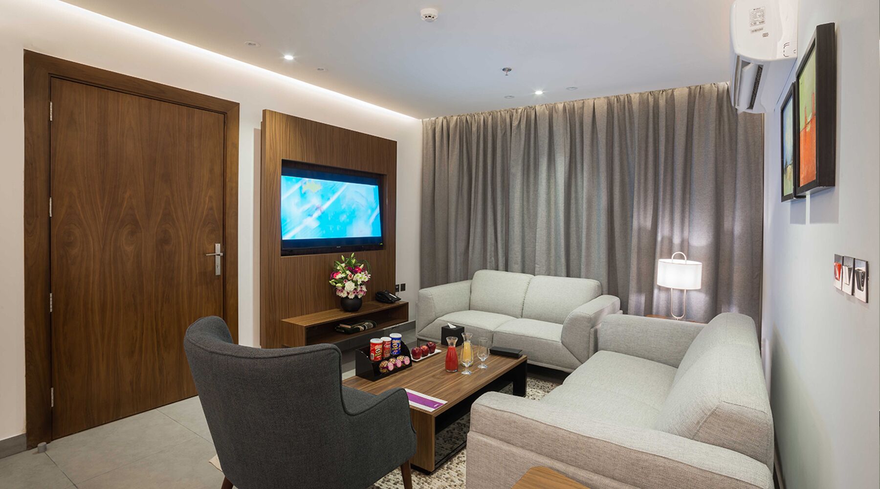 Ett modernt hotellrum med en grå soffa, ett träskåp med en tv, ett litet bord med snacks och ett stort fönster med gardiner.