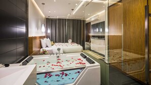 A modern hotel room with a large bed, a separate sitting area, a bathtub with rose petals, and a glass partition. - Boudl Al Wadi (Riyadh)