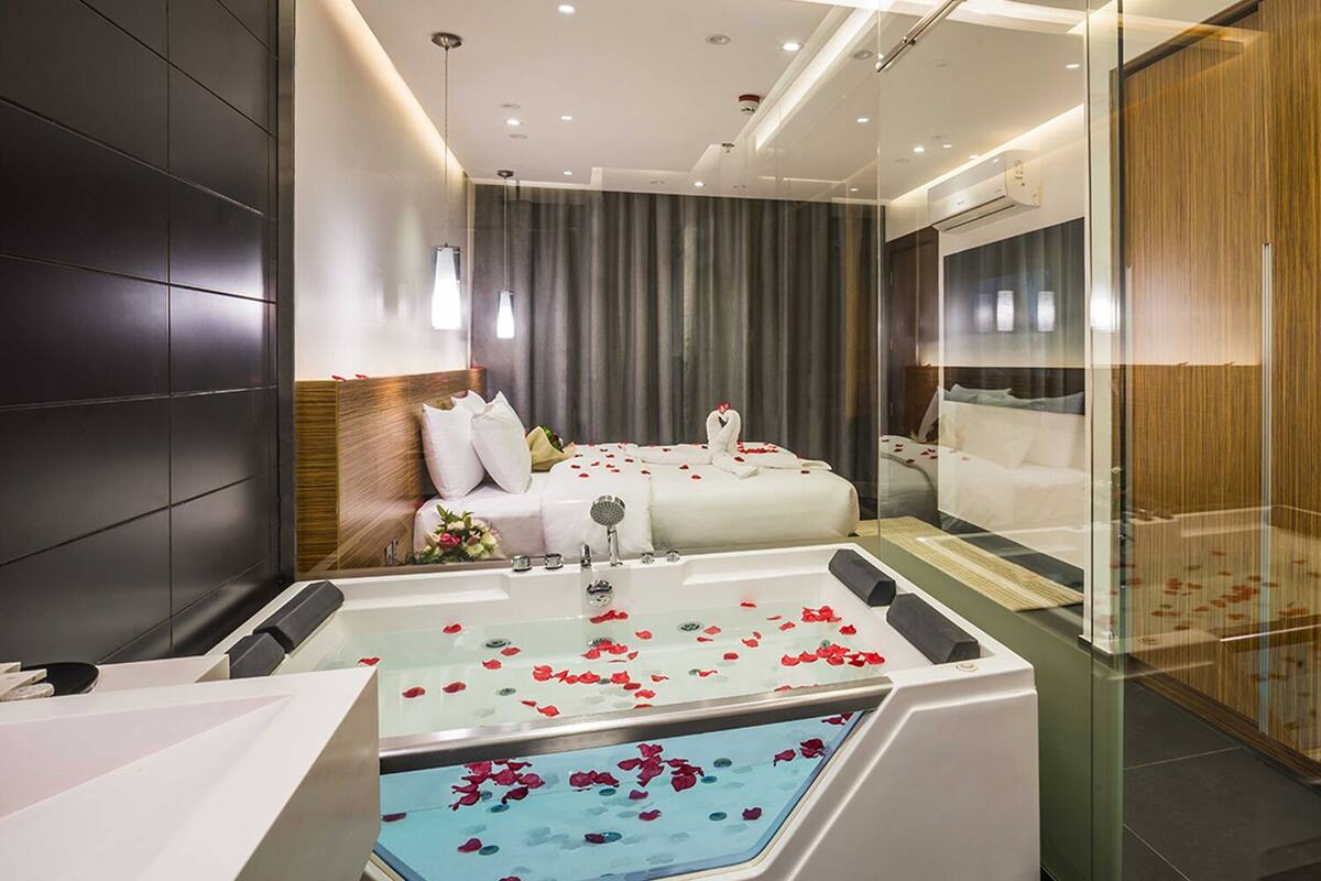 Una habitación de hotel moderna con una cama grande, una zona de estar separada, una bañera con pétalos de rosa y un separador de vidrio.