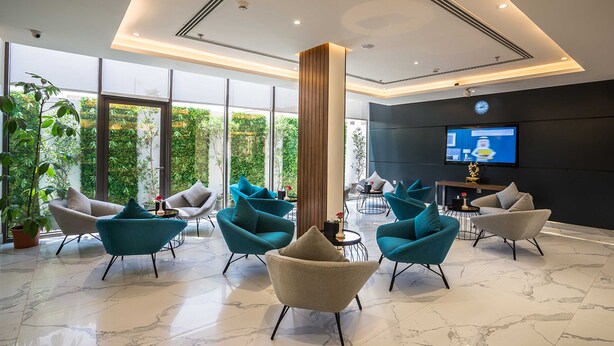 A modern hotel lobby with a large glass wall, multiple seating areas with teal and grey chairs, a central wooden column, and a flat-screen TV on the wall. - Boudl Al Wadi (Riyadh)