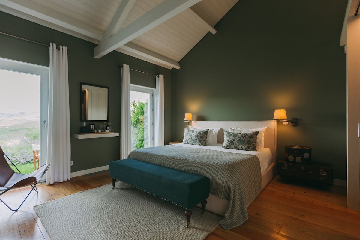 Un dormitorio con una cama grande, un banco, una silla, una ventana con cortinas y vistas a zonas verdes.