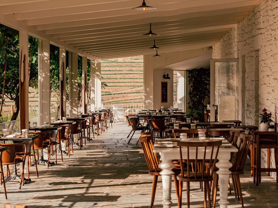 A spacious indoor dining area with long tables and chairs, large windows, and a stone floor.