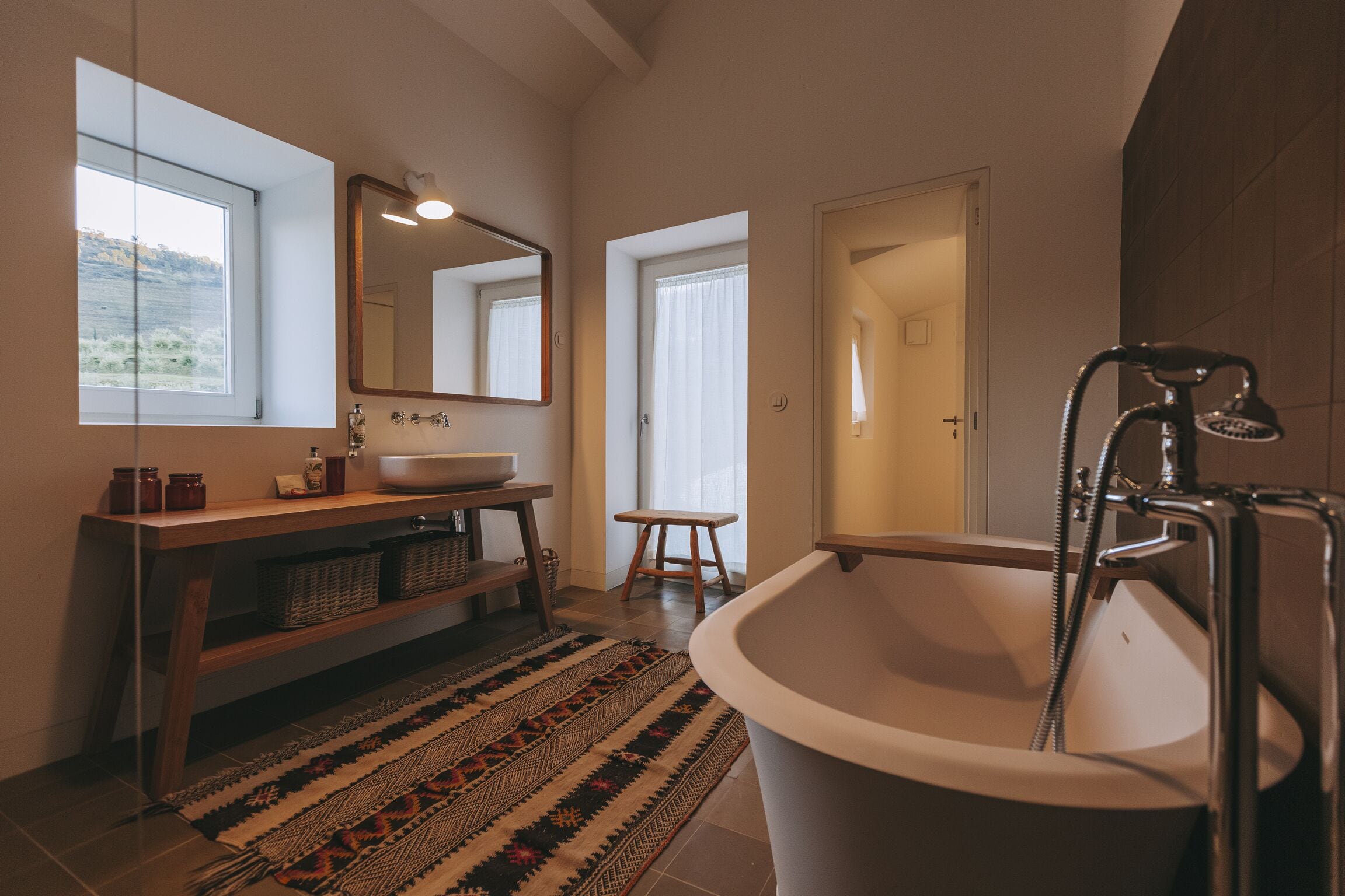 Un baño moderno con una bañera independiente, un espejo grande, un mueble de tocador de madera y una alfombra estampada.