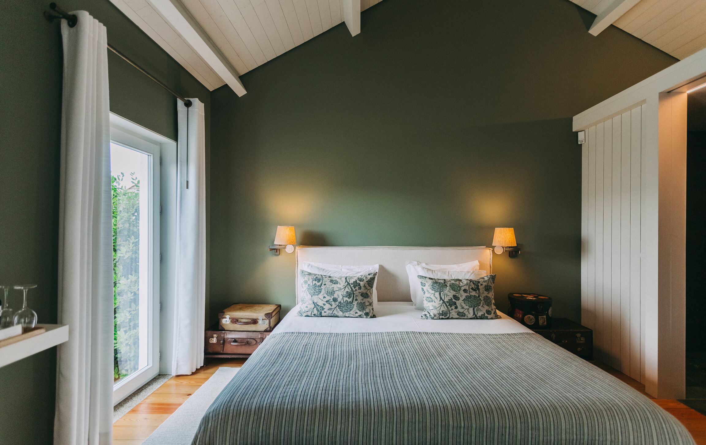 Un dormitorio con una cama grande, dos lámparas de noche, una ventana con cortinas, una mesita de noche y una maleta.