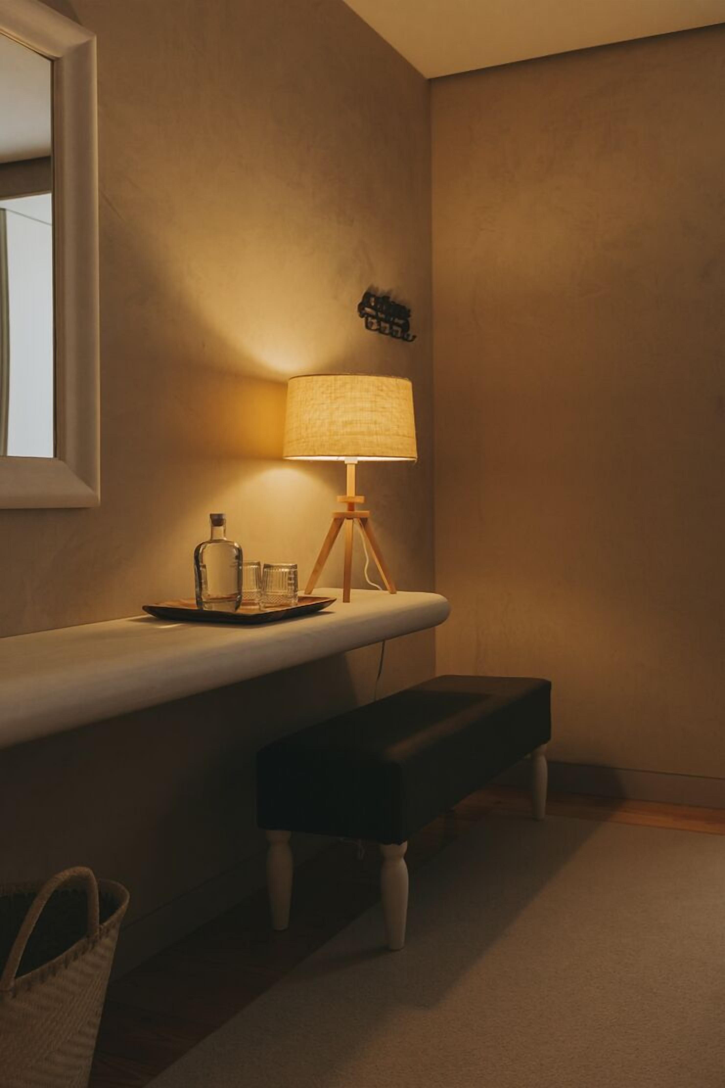 A dimly lit room with a bedside table, a lamp, a bench, and a small wall-mounted shelf.