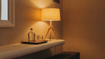 Una habitación tenuemente iluminada con una mesita de noche, una lámpara, un banco y un pequeño estante en la pared.