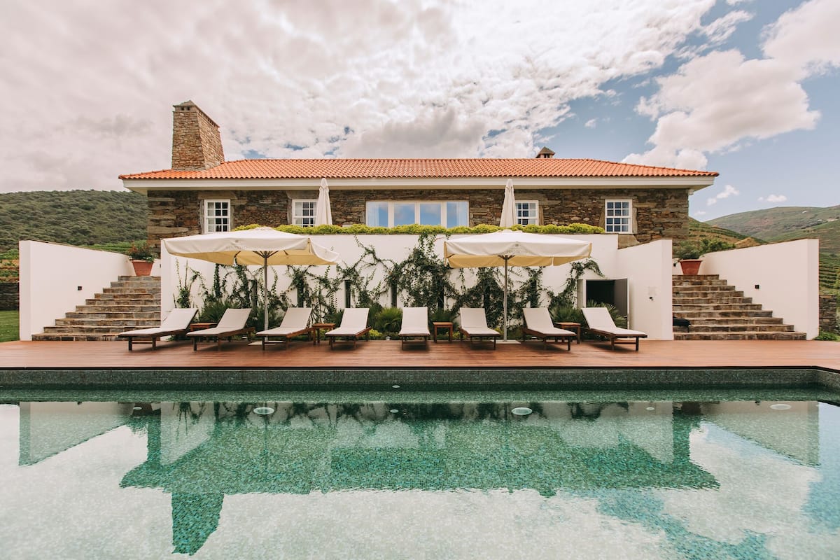 Un edificio de piedra con techo de tejas, piscina y mobiliario exterior con sombrillas.