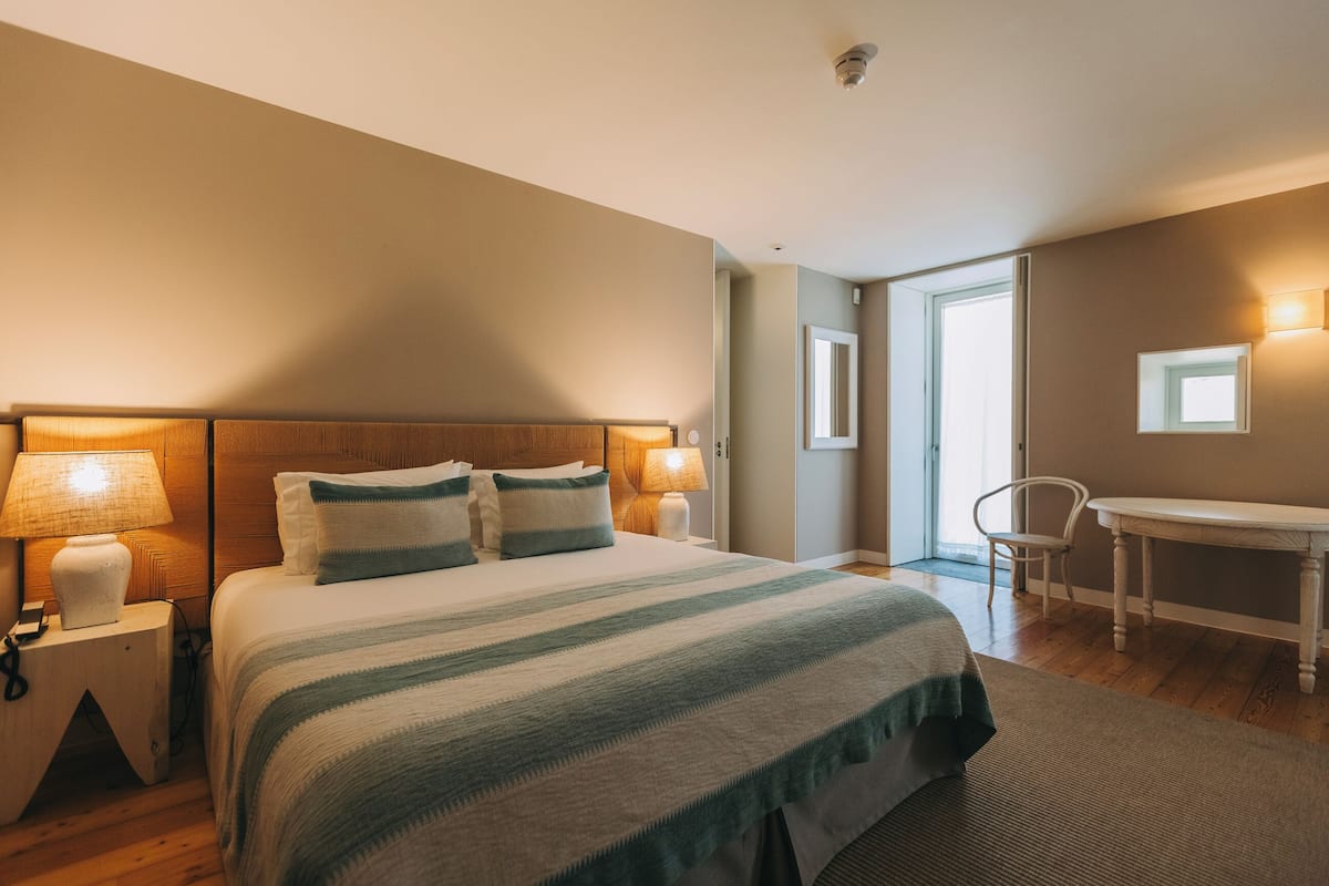 Una habitación de hotel con una cama grande, lámparas de noche, una mesita y una silla.