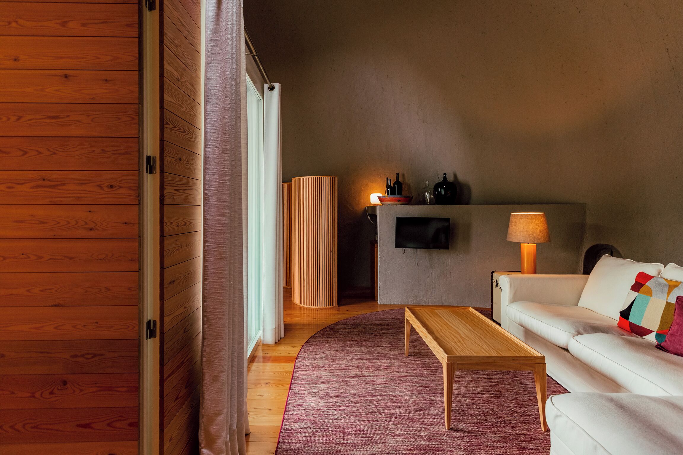 Un salón moderno con suelo de madera, un sofá blanco, una mesa de centro de madera y una alfombra roja.