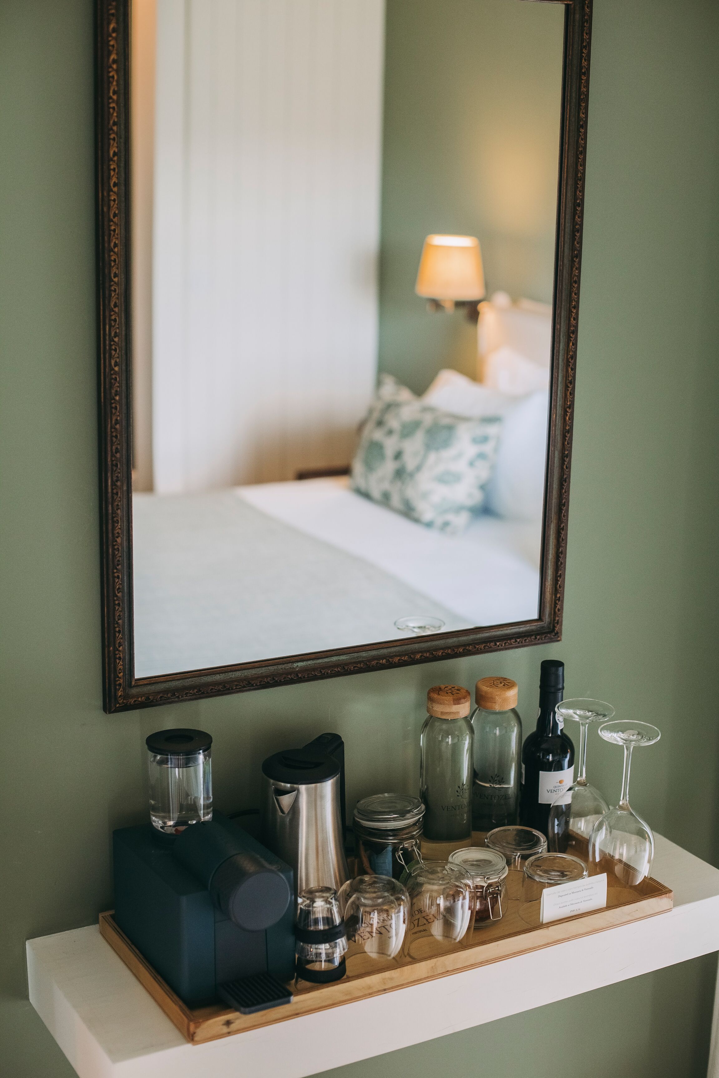 Quarto de hotel com cama, mesa de cabeceira, espelho e um apoio com diversos objetos, incluindo uma cafeteira, potes e copos.