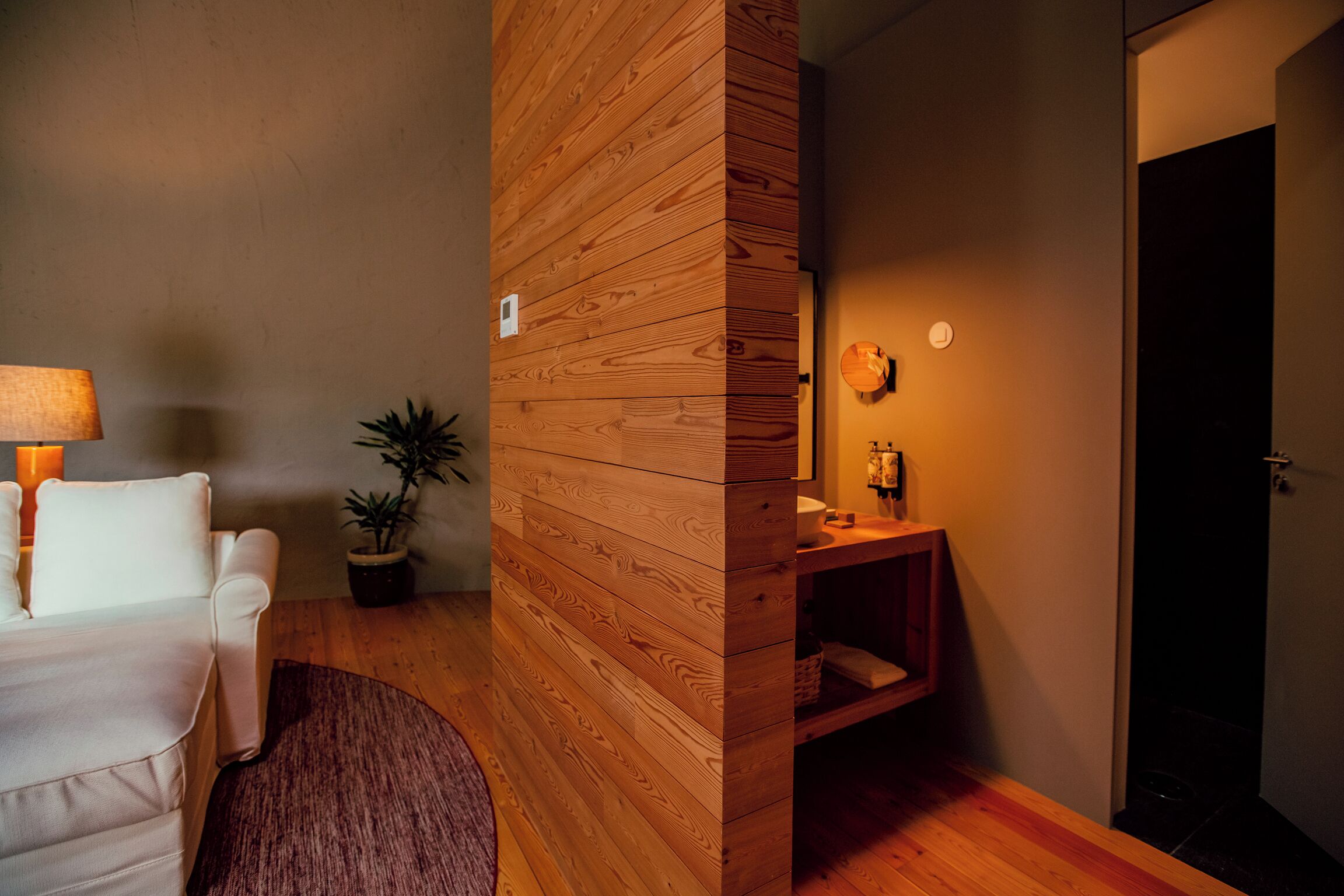 A modern room with a wooden accent wall, a white sofa, a small table with a lamp, a potted plant, and a wooden cabinet with a mirror.