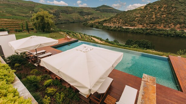 Zona de piscina con terraza de madera y sombrillas blancas, con vistas a un río y un paisaje montañoso.