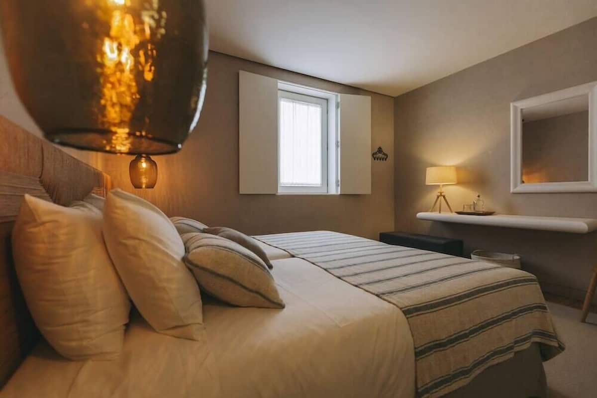 Un dormitorio con cama, almohadas, lámpara de noche, ventana y estante en la pared.