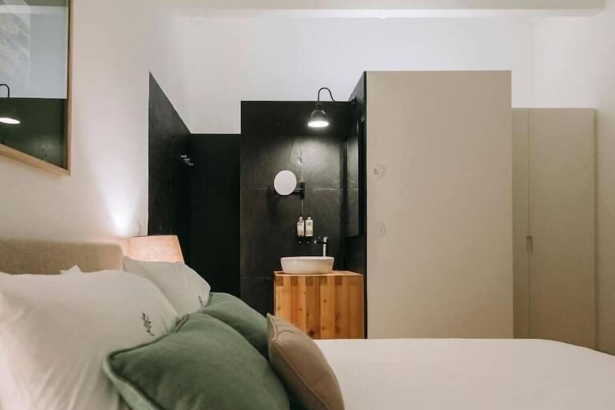 Un dormitorio con cama, almohadas, mesita de noche, espejo, armario y baño con lavamanos y espejo.