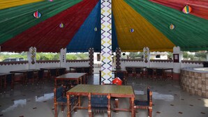 A covered outdoor dining area with multiple wooden tables and chairs, a central pillar with a patterned design, and colorful drapes. - Rann Village Resort (Bhuj)