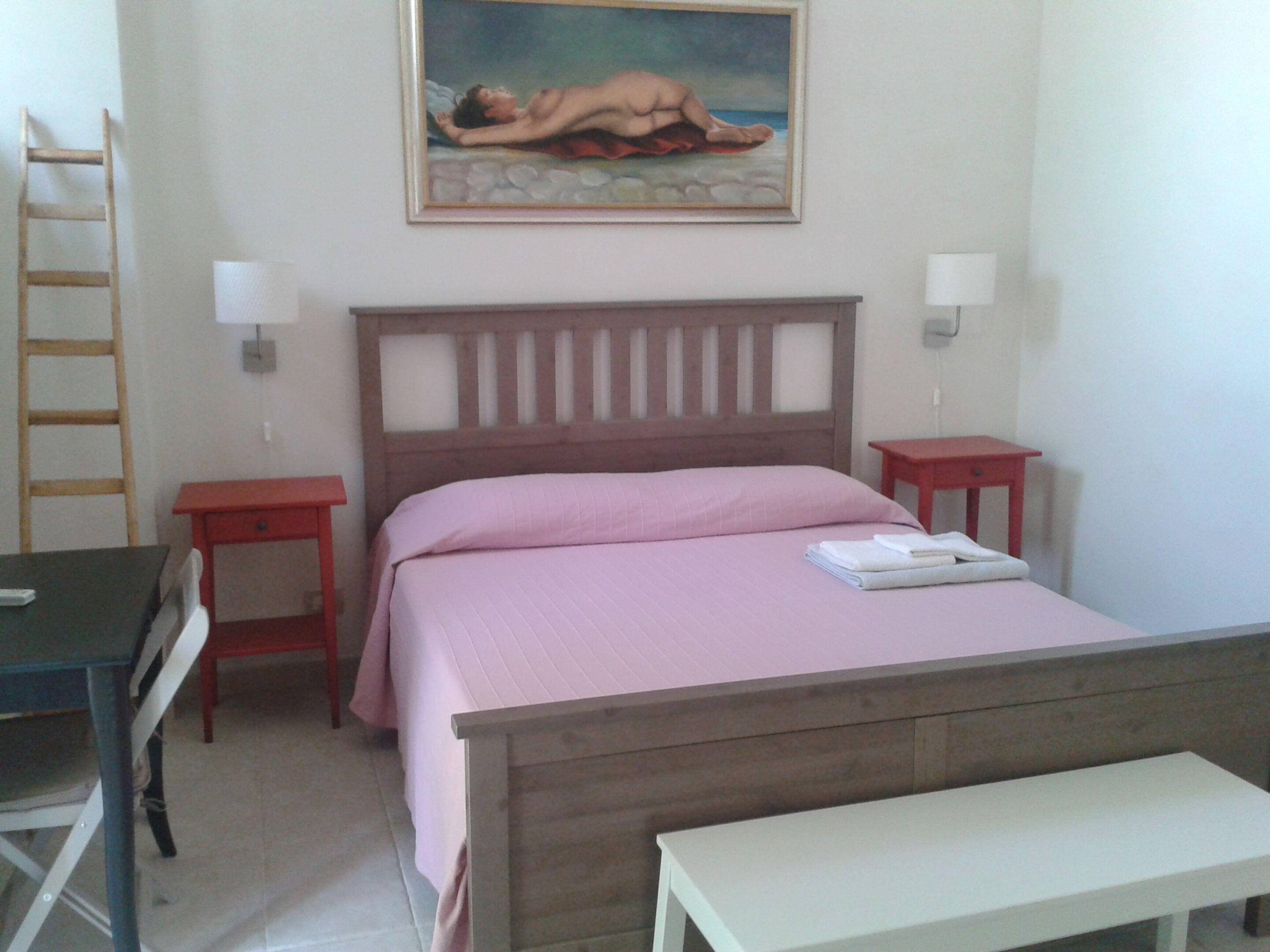 A bedroom with a wooden bed, pink bedding, two bedside tables, a nightstand, a desk, and a ladder against the wall.