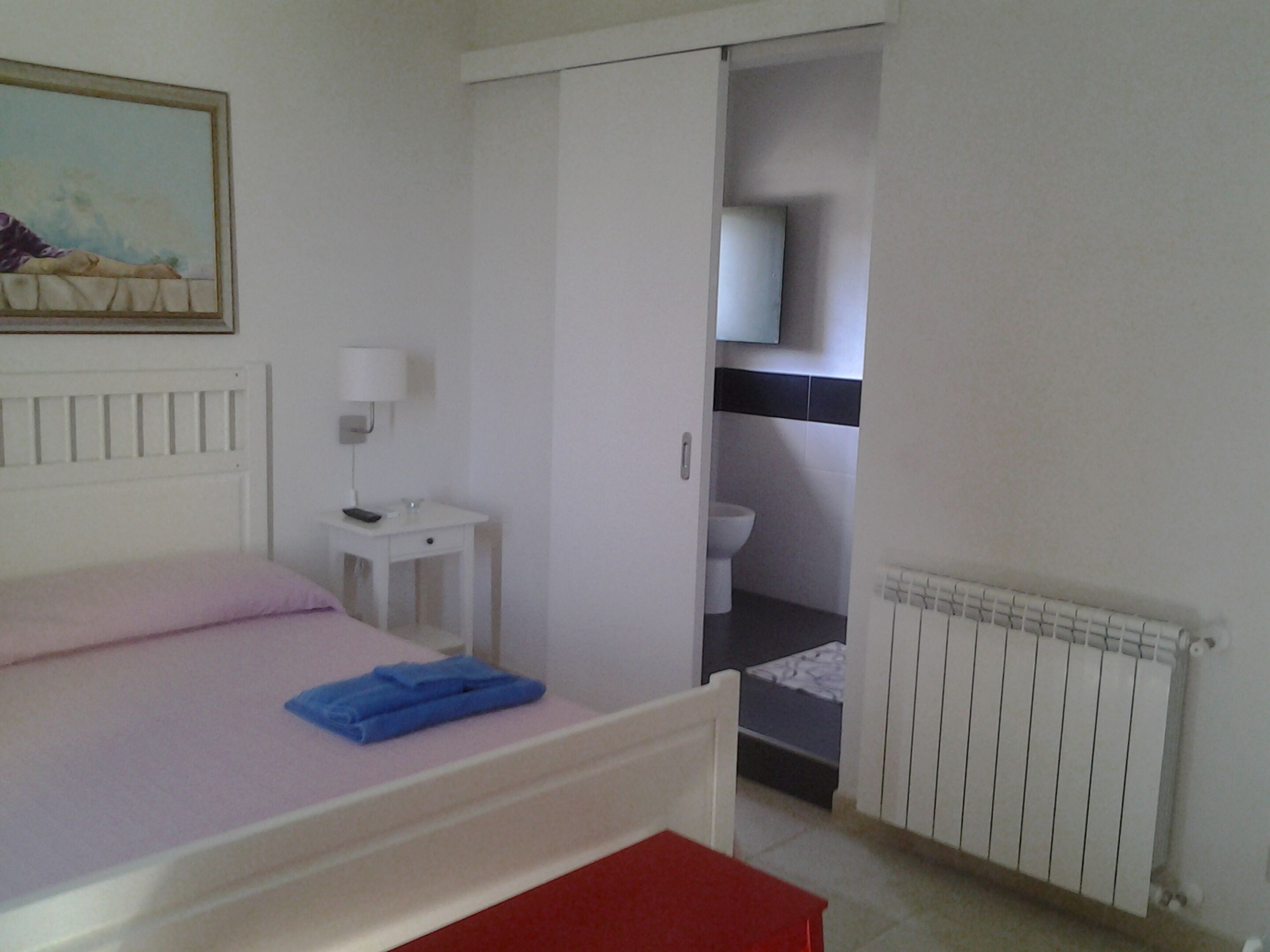 A bedroom with a bed, a bedside table with a lamp, a small table with a remote, a red bench, and a bathroom visible through an open door.
