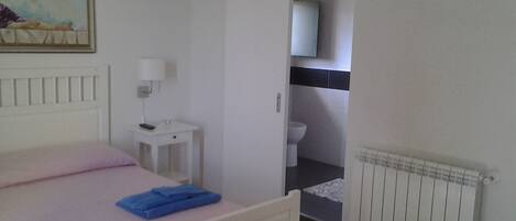 A bedroom with a bed, a bedside table with a lamp, a small table with a remote, a red bench, and a bathroom visible through an open door.