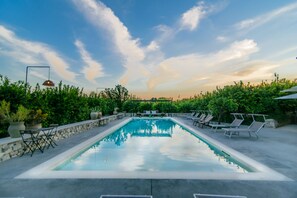 A rectangular swimming pool with lounge chairs and tables on either side, surrounded by greenery and a stone wall. - Tenuta Don Paolino (Avola)