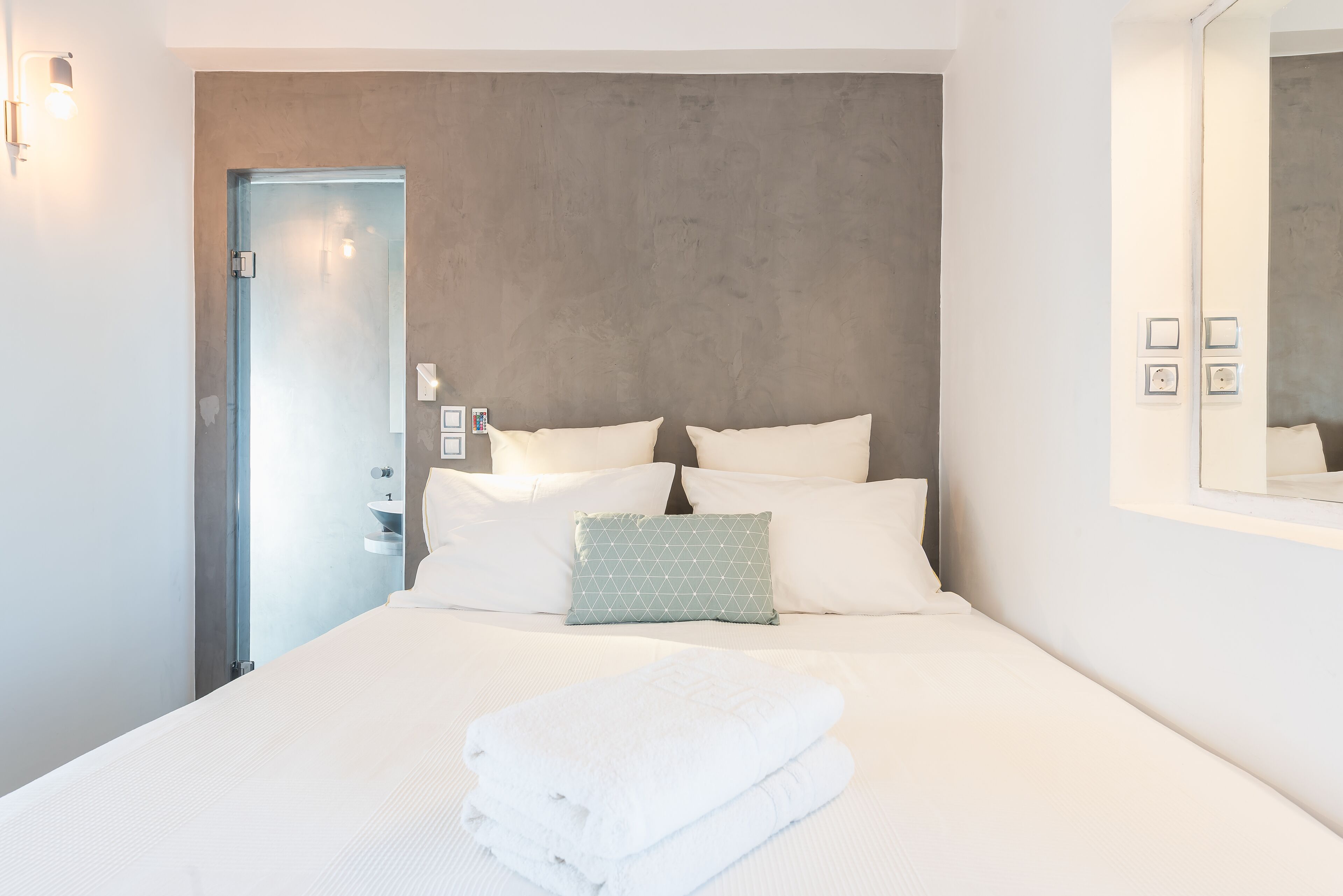 A neatly made bed with white linens and a folded towel, a grey textured wall behind, and a door to the left.