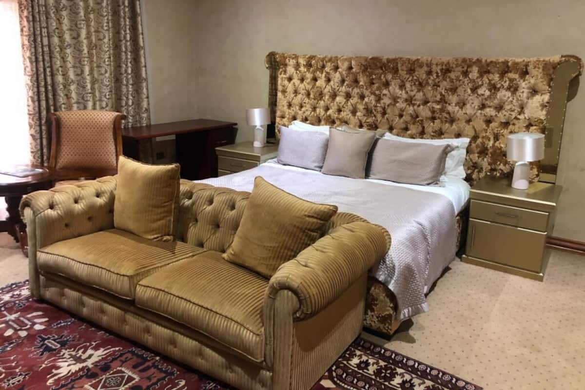 A hotel room with a tufted headboard bed, a brown sofa, a patterned rug, and a wooden nightstand with a lamp.