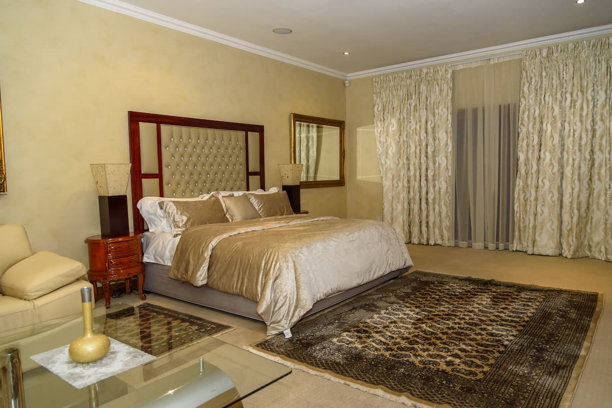 A bedroom with a bed, a nightstand, a lamp, a sofa, and a large window with curtains.