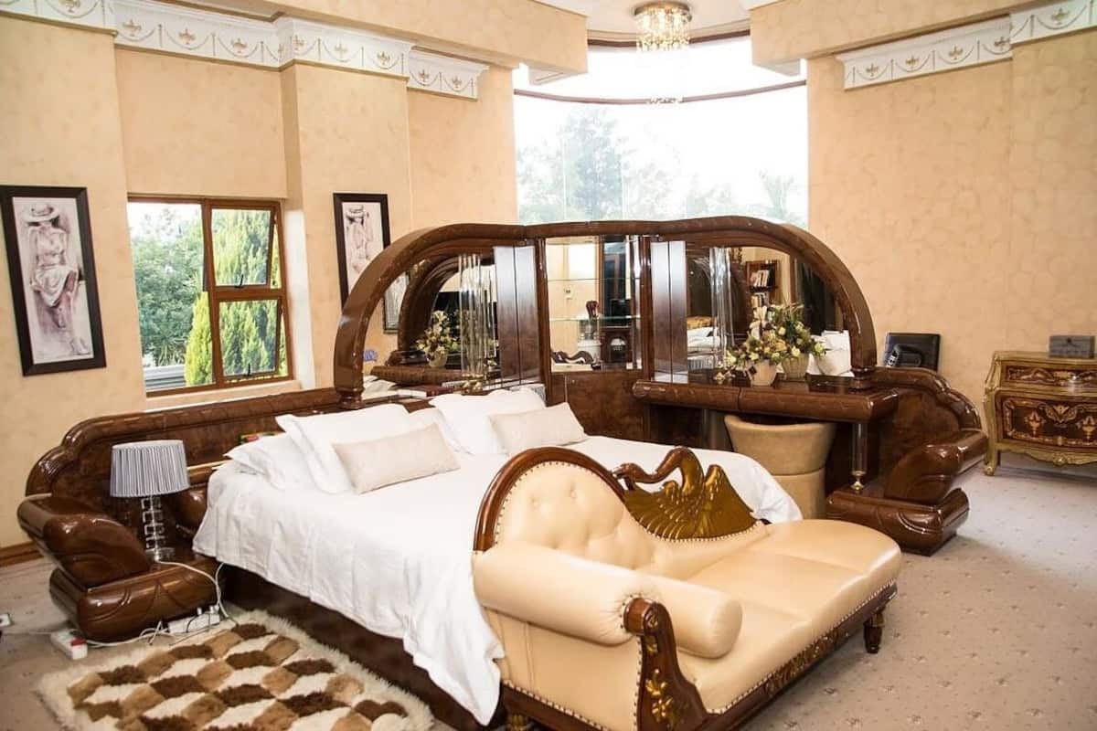 A luxurious bedroom with a large bed, a chaise lounge, a vanity with a mirror, and a dresser.