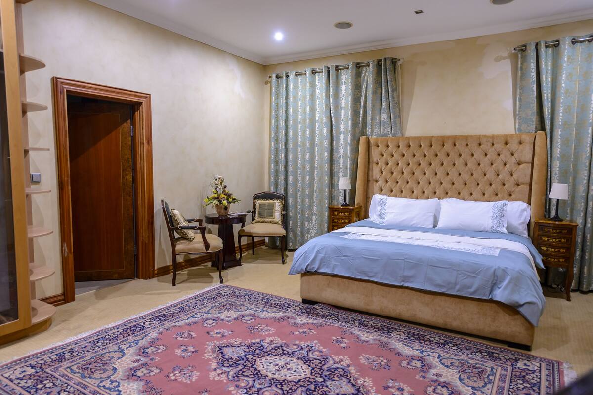 A bedroom with a large bed, a chair, a small table with a vase, and a wooden wardrobe.
