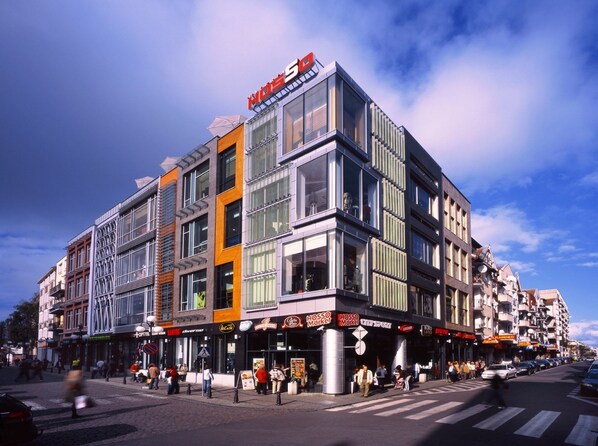 A modern hotel with a prominent 'HOTEL S' sign, featuring a mix of glass and brick facades, and a variety of shops on the ground floor.