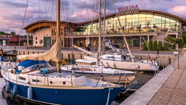 Un puerto deportivo con varios veleros amarrados, un edificio con la inscripción "MARINA SOLNA KOLOBRZEG" al fondo y un paseo marítimo.