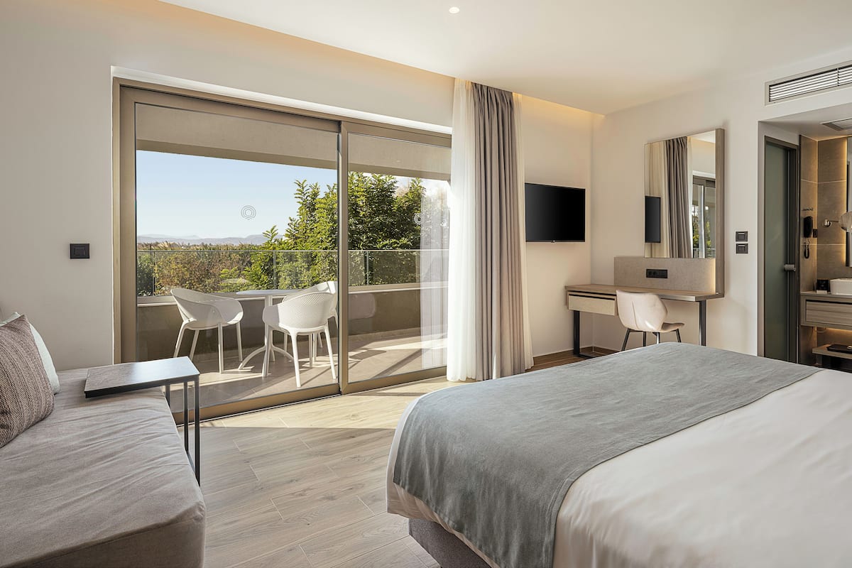 A modern hotel room with a large bed, a desk, a chair, a balcony with a view, and a bathroom with a mirror and sink.