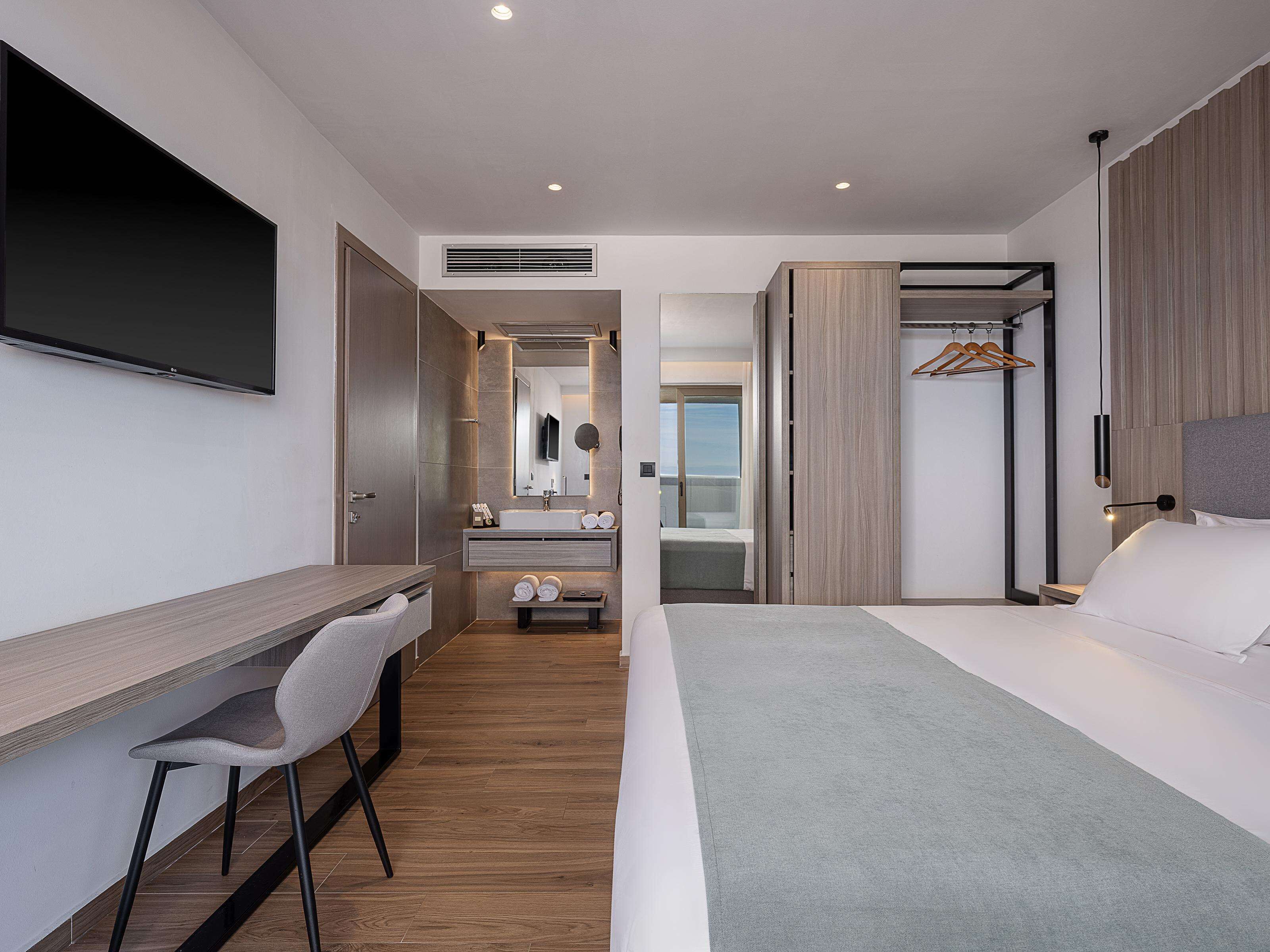 Een moderne hotelkamer met een bureau, stoel, bed en een inloopkast met spiegel en hanggedeelte.