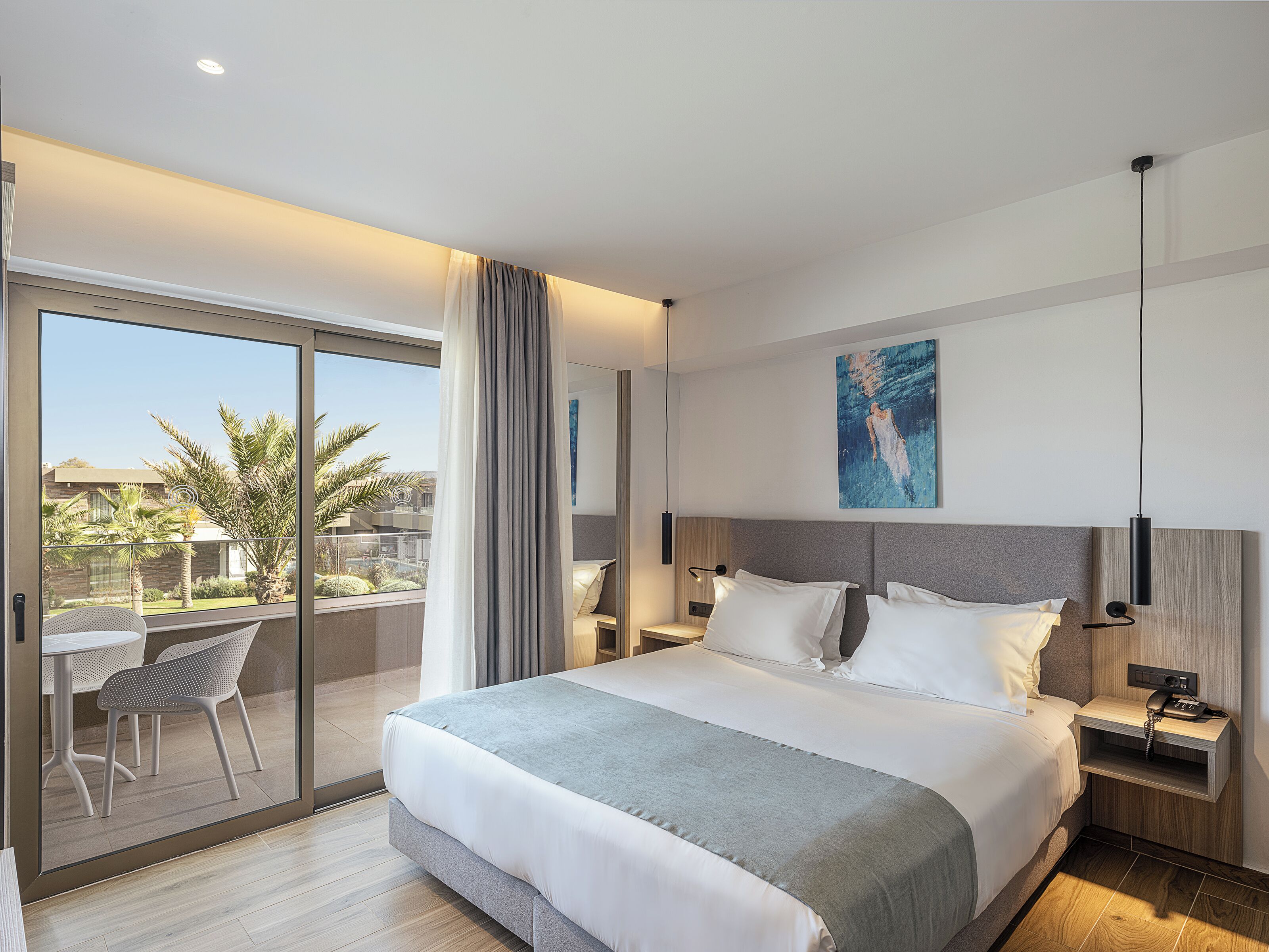 Ein modernes Hotelzimmer mit einem großen Bett, einem Balkon mit Tisch und Stühlen und Blick auf Palmen.