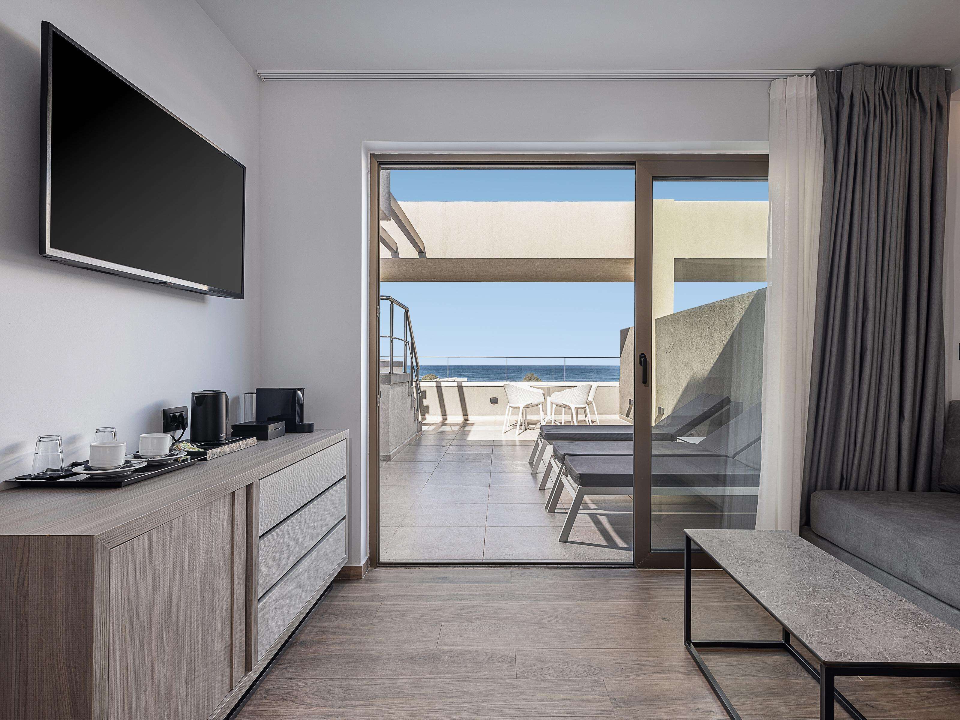 Ein modernes Hotelzimmer mit Fernseher, einem Holzschrank, einem Balkon mit Meerblick und einer Sitzecke mit Sofa und Tisch.