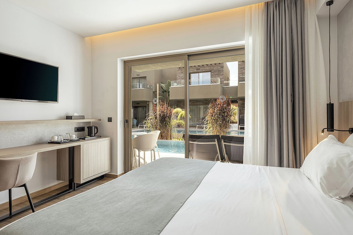 A hotel room with a bed, a desk, a TV, a view of a pool and outdoor seating, and a sliding door to the balcony.