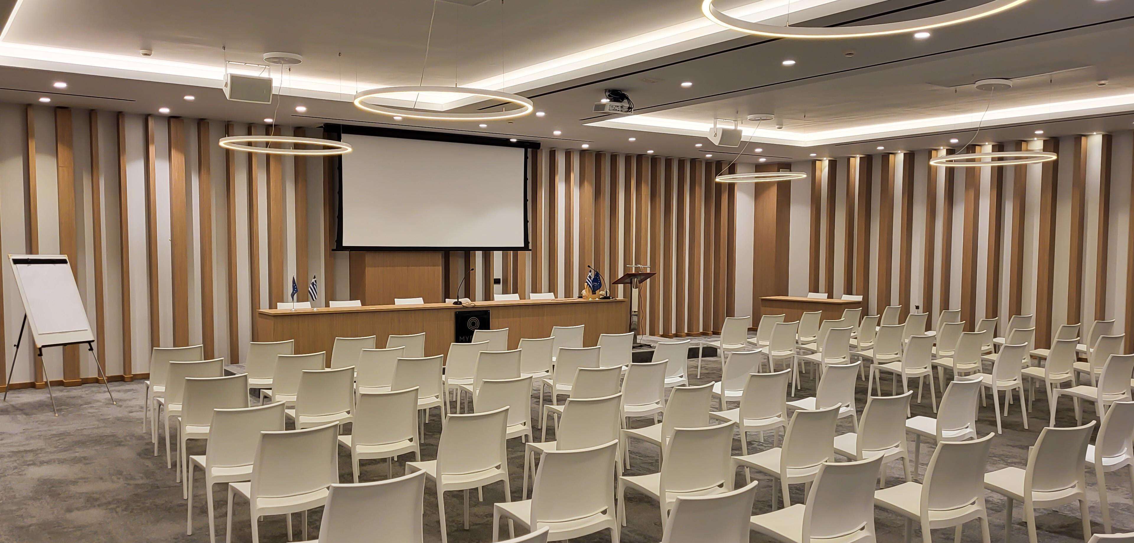 Uma sala de conferência com um púlpito, uma tela grande e fileiras de cadeiras brancas.