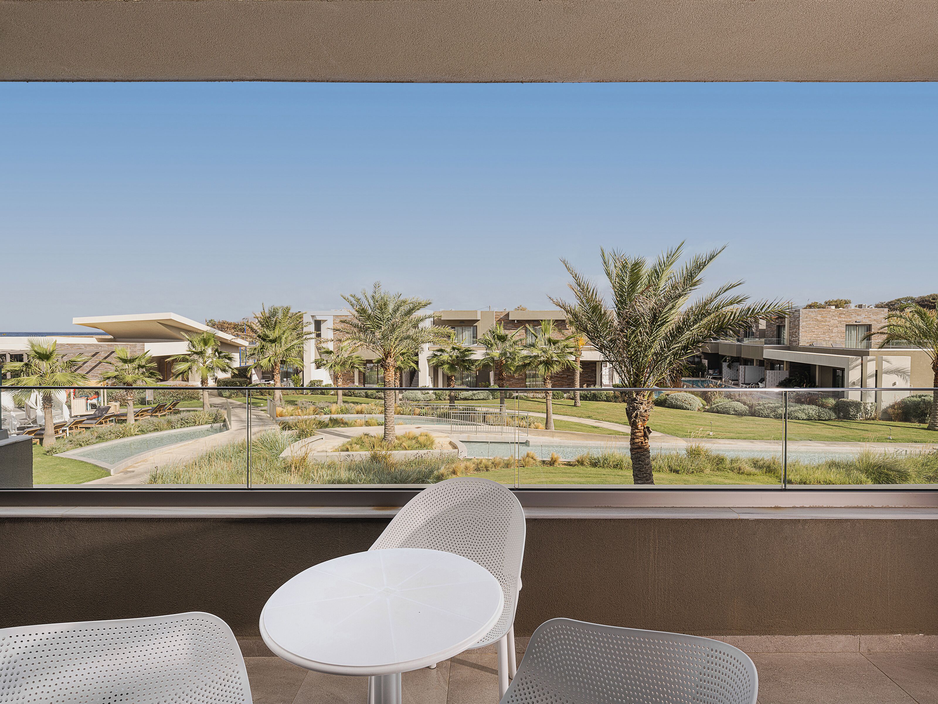A balcony with two white tables and chairs, overlooking a landscaped area with palm trees and a pool.